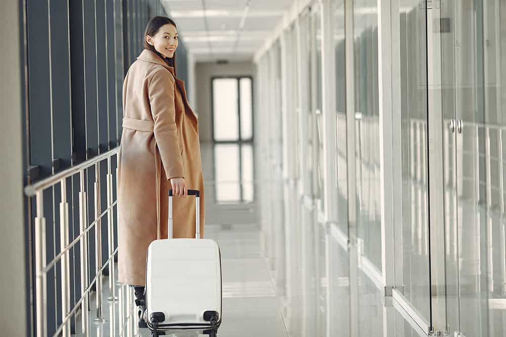 Woman Dressed In A Brown Trench Carrying A White Suitcase In The Airport For A Business Trip