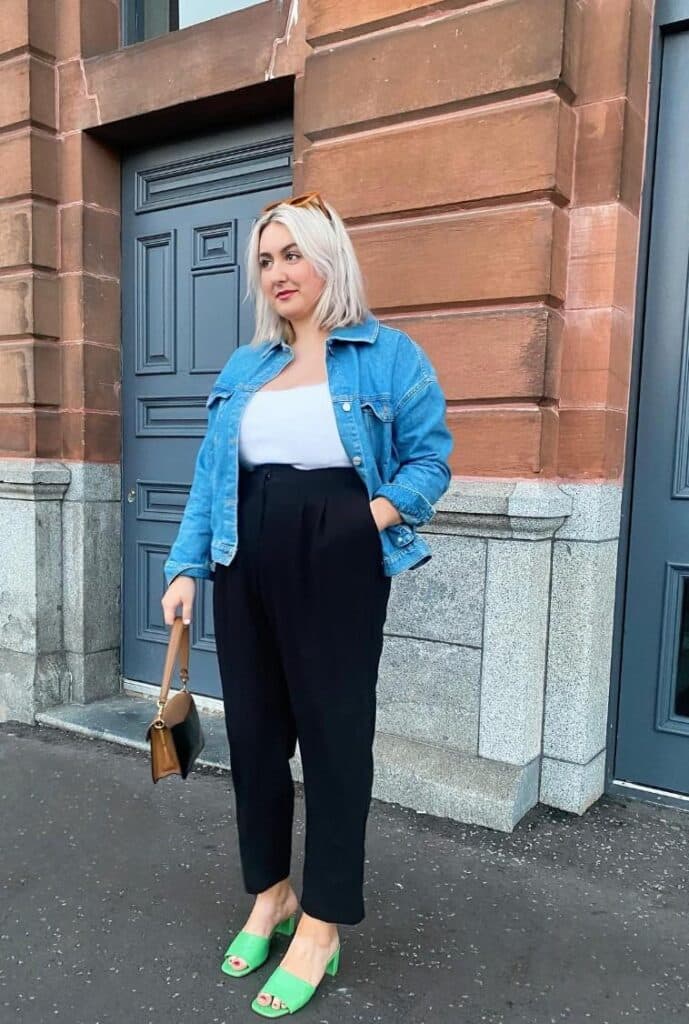 Mid-size woman in a business casual outfit consisting of a white blouse, black capri pants, a light jean jacket, and emerald green workwear shoes.